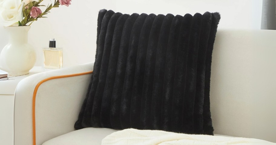 a black color ribbed faux fur pillow on a tan sofa next to a side table with a vase of flowers on it