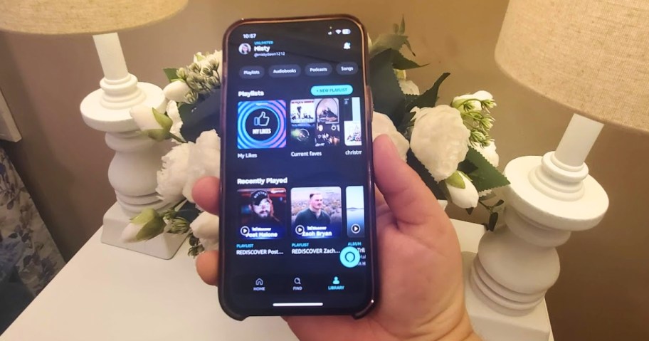 hand holding an iPhone with the Amazon Music Unlimited app open showing playlists, recent plays, and more, vase of faux white flowers and lamps on a table in the background