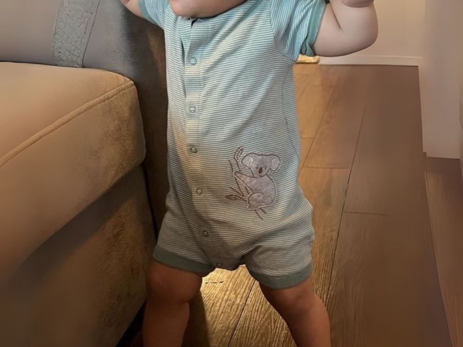 baby standing up next to couch wearing a green striped romper with a koala on it