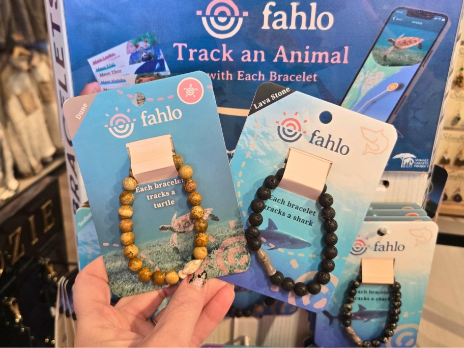 hand holding Fahlo animal tracking beaded bracelets on product cards, display with more bracelets behind it