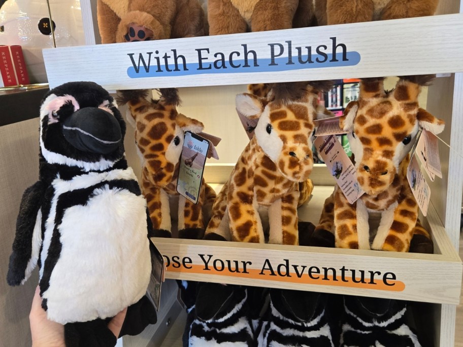 hand holding a penguin plush next to a display with more plush animals including Giraffes