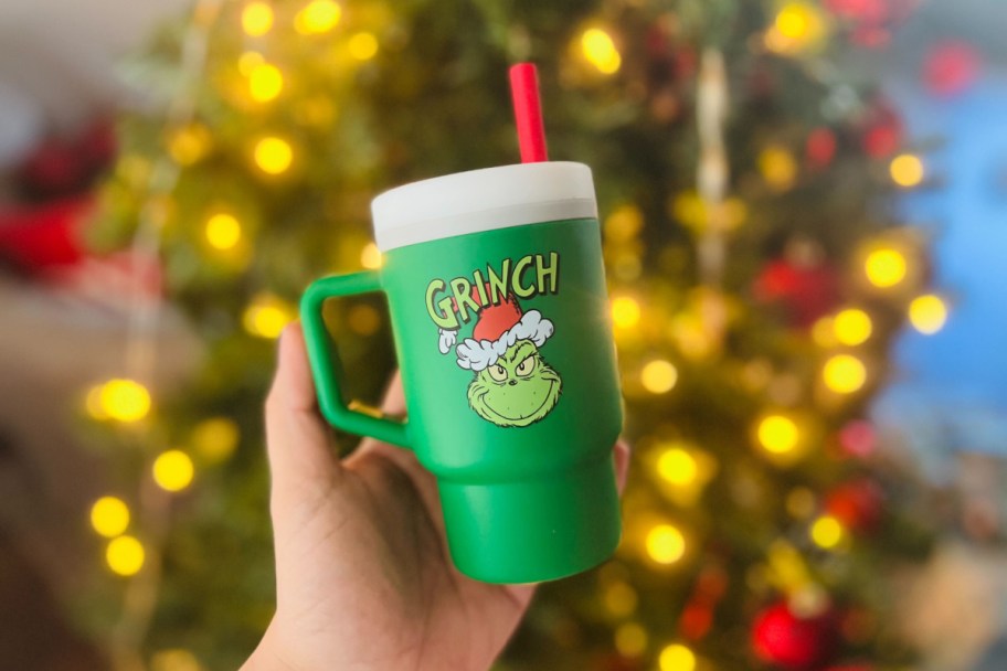 hand holding an infantino my first tumbler grinch cup in front of a lit christmas tree