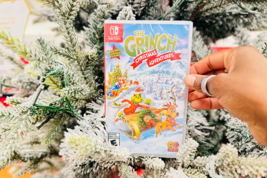 hand holding a grinch nintendo switch game in front of a flocked christmas tree