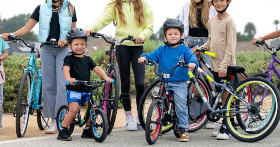 little and big kids sitting on or standing by bicycles in various sizes and colors, all are wearing helmets