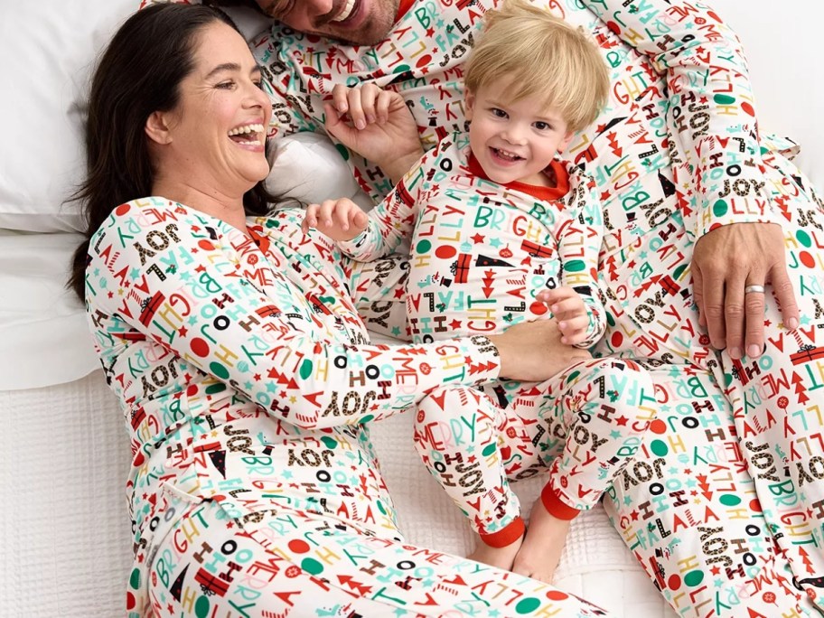 mom, dad, and toddler wearing matching Christmas pajamas that are cream and have the words various holiday phrases and words on them in red, green, blue, and yellow colors