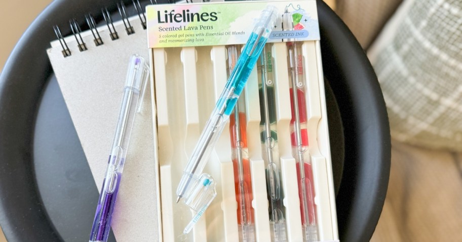 a pack of Lifeline Scented Lava Gel pens in the box, a blue pen on top of the box, writing pad underneath the box on a side table