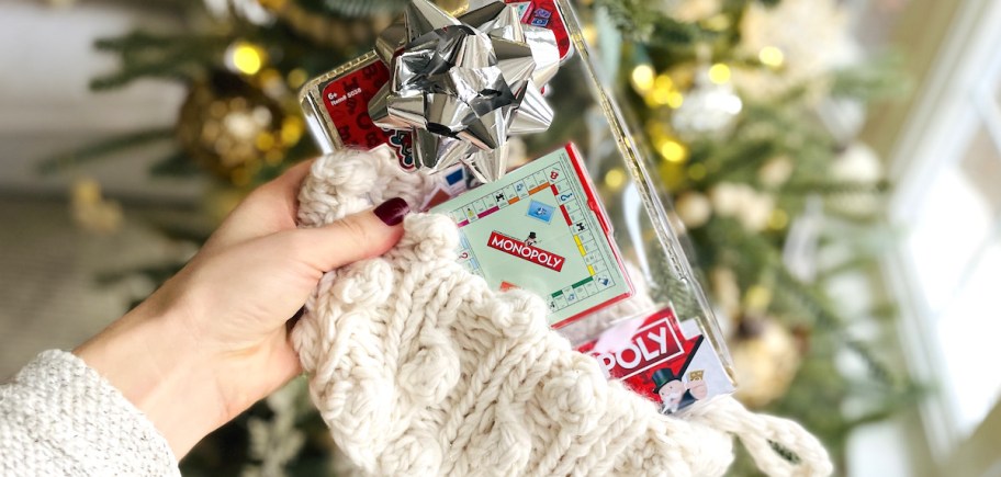 hand holding a white knit stocking with mini monopoly game inside