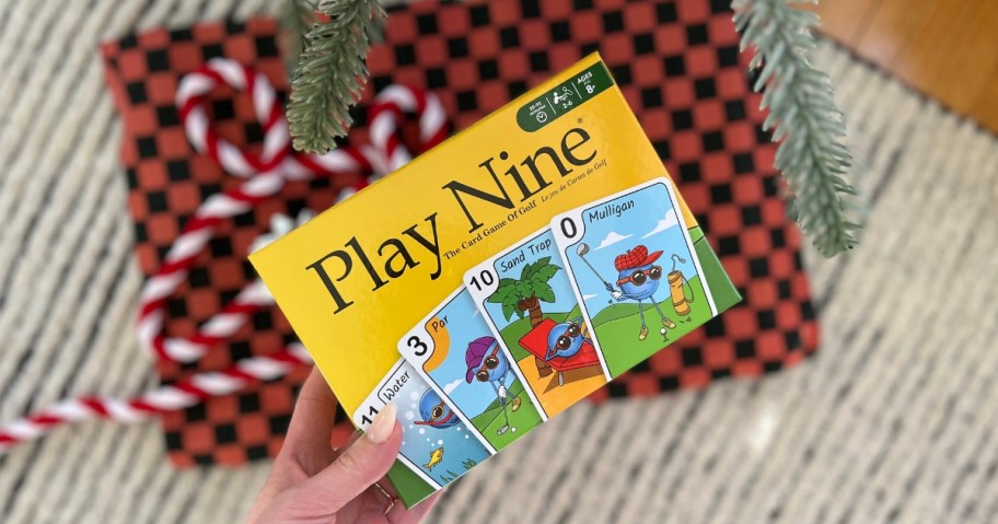 hand holding a yellow box for Play Nine gold card game, Christmas present and tree behind it