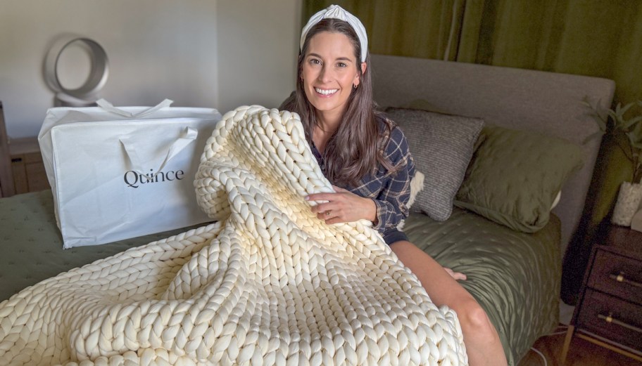 woman sitting on bed holding cream weighted blanket next to quince bag