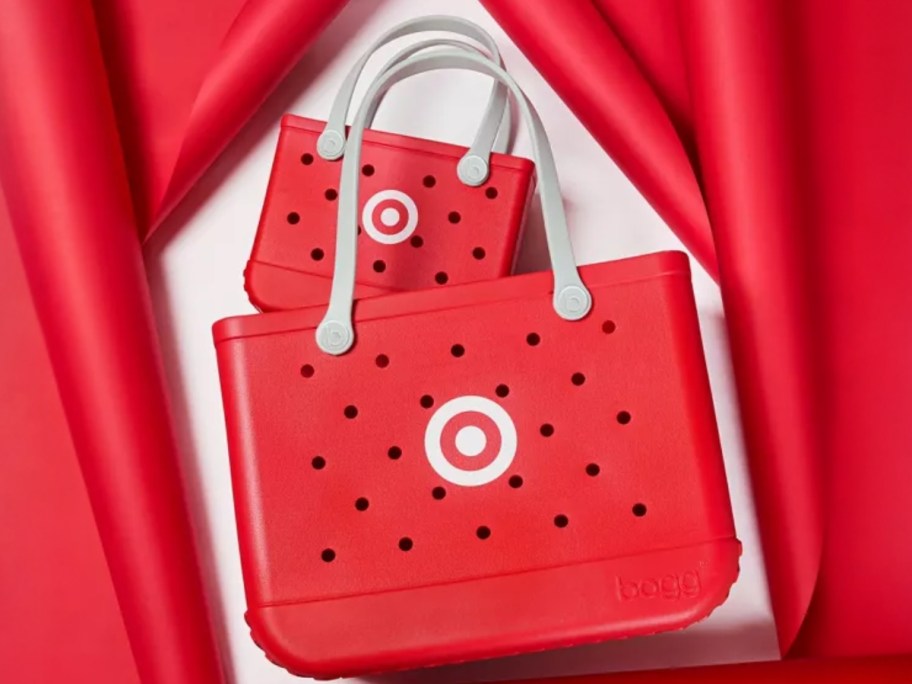 2 Bogg Bags (plastic tote bags with holes) in red with white handles and the Target bullseye logo on them laying on a back background with red curtains on each side