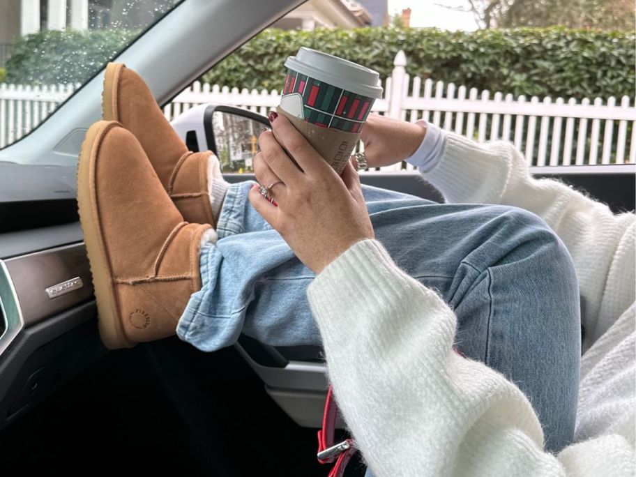 Woman sitting in the passenger seat of a car holding a cup of coffee with her feet up on the dash wearing Dearfoams Fireside Riverland Boots