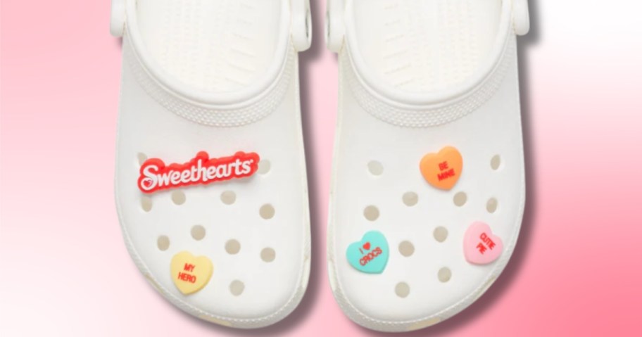 a pair of white Crocs clog shoes with Sweethearts Jibbitz shoe charms that look like candy hearts with phrases on them