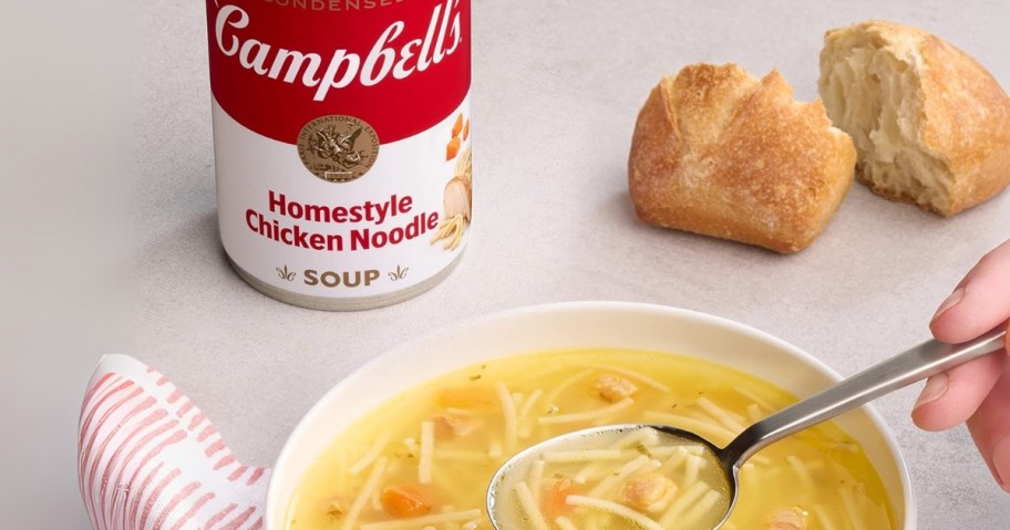 a bowl of chicken noodle soup with a person's hand holding a spoon in it, with a can of Campbell's Homestyle Chicken Noodle Soup and some bread in the background