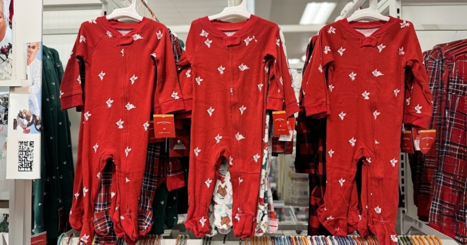 3 baby footie onesie union suit pajamas hanging from a store display, they are red with the Target Bullseye white dog on them