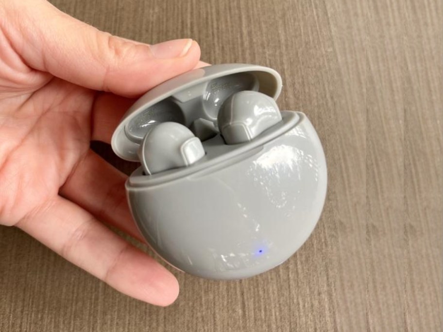 hand holding a pair of grey earbuds in a grey charging case, woodgrain tabletop behind it