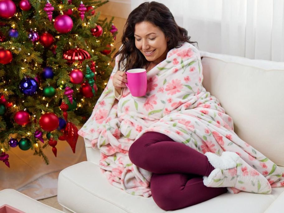 Woman wrapped in a throw blanket wearing matching booties sitting on a couch drinking from a mug