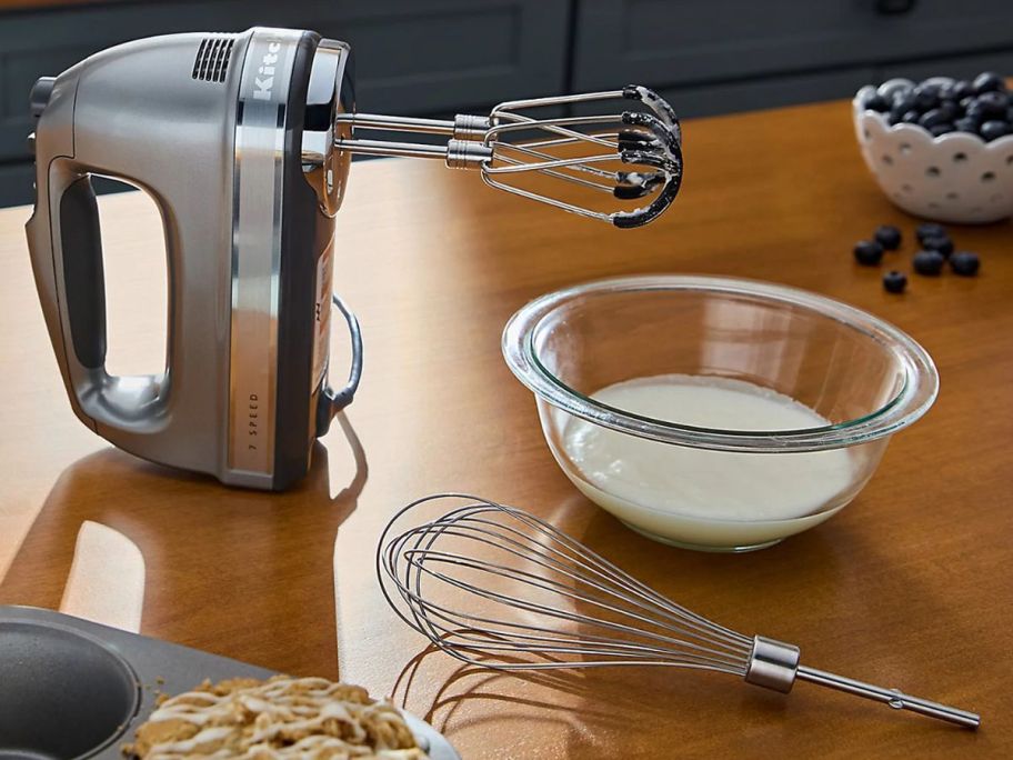 A silver Kitchenaid Mixer with flex edge beaters next to a bowl of white liquid and a whisk attachment