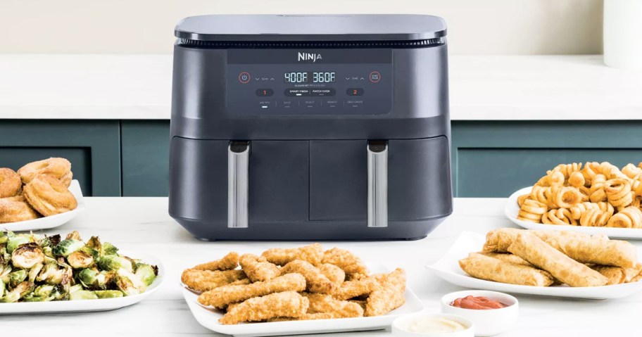 grey air fryer on counter with plates of food around it