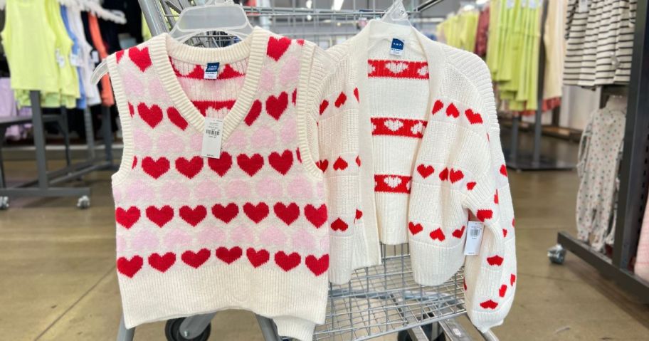 A heart sweater vest and cardigan from Old Navy hanging on a shopping cart