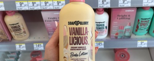 A person holds a 500 ml bottle of "Soap & Glory Vanilla-Licious" body lotion with a creamy vanilla scent in front of a store shelf filled with colorful skincare products.