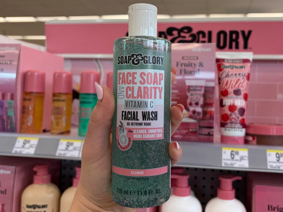 A hand holds a bottle of Soap & Glory Face Soap and Clarity Vitamin C Facial Wash in a store. The shelf behind displays various pink skincare products.