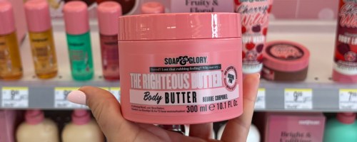 A hand holds a pink container of "Soap & Glory The Righteous Butter" body butter, 300ml. The background shows a store shelf with various pink cosmetic products.