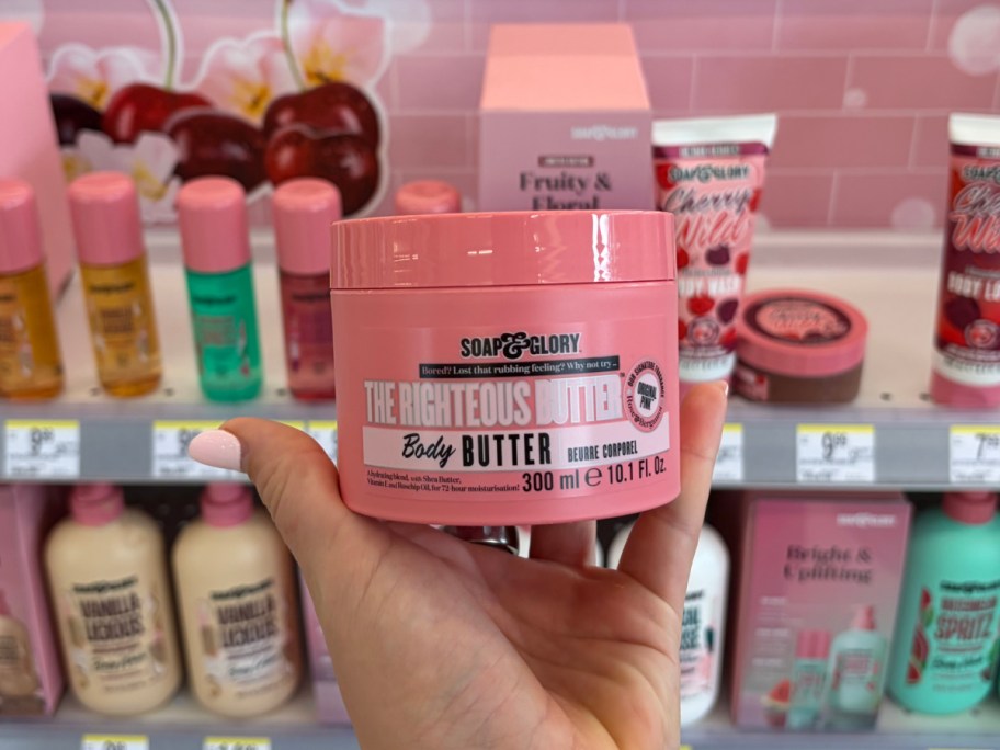A hand holds a pink container of "Soap & Glory The Righteous Butter" body butter, 300ml. The background shows a store shelf with various pink cosmetic products.