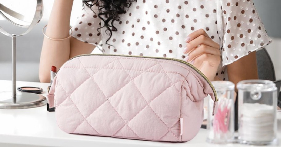 A pink quilted cosmetic bag sitting on a vanity table next to various beauty products.