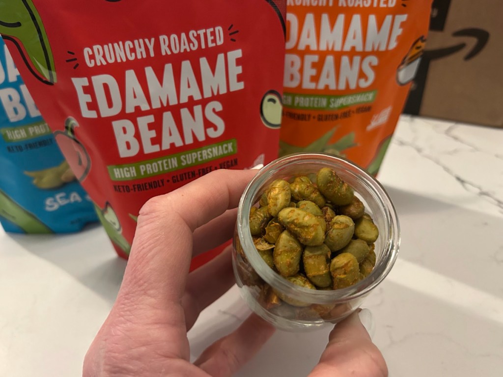 Small glass jar of roasted edamame beans in front of several different snack bags