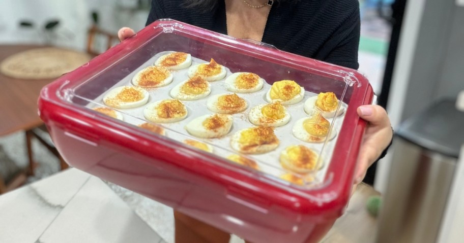 woman holding a dark red color food container with a clear top, you can see a tray of devilled eggs inside