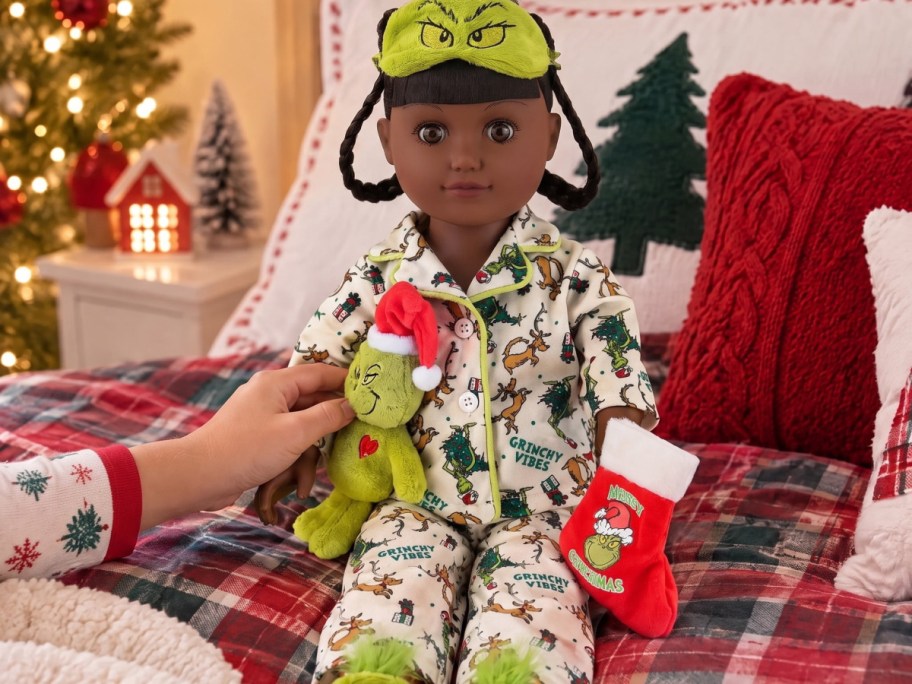 A fashion doll wearing Grinch pajamas and a matching eye mask sits on a festive bed holding a small Grinch plush toy and a miniature Christmas stocking.