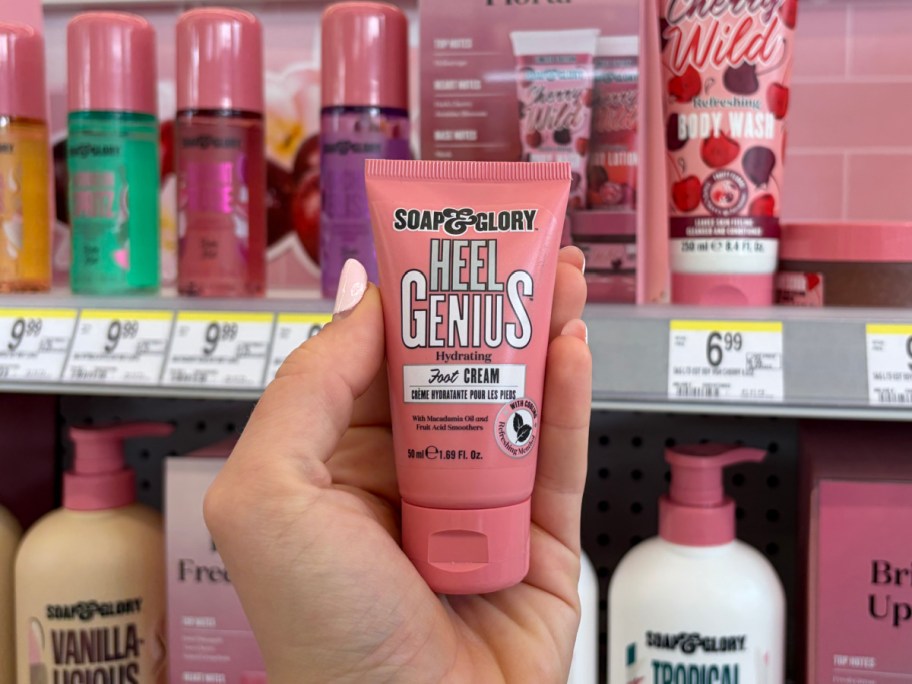A hand holding a pink tube of "Soap & Glory Heel Genius Foot Cream" in a store. The shelf in the background displays various colorful skincare products.