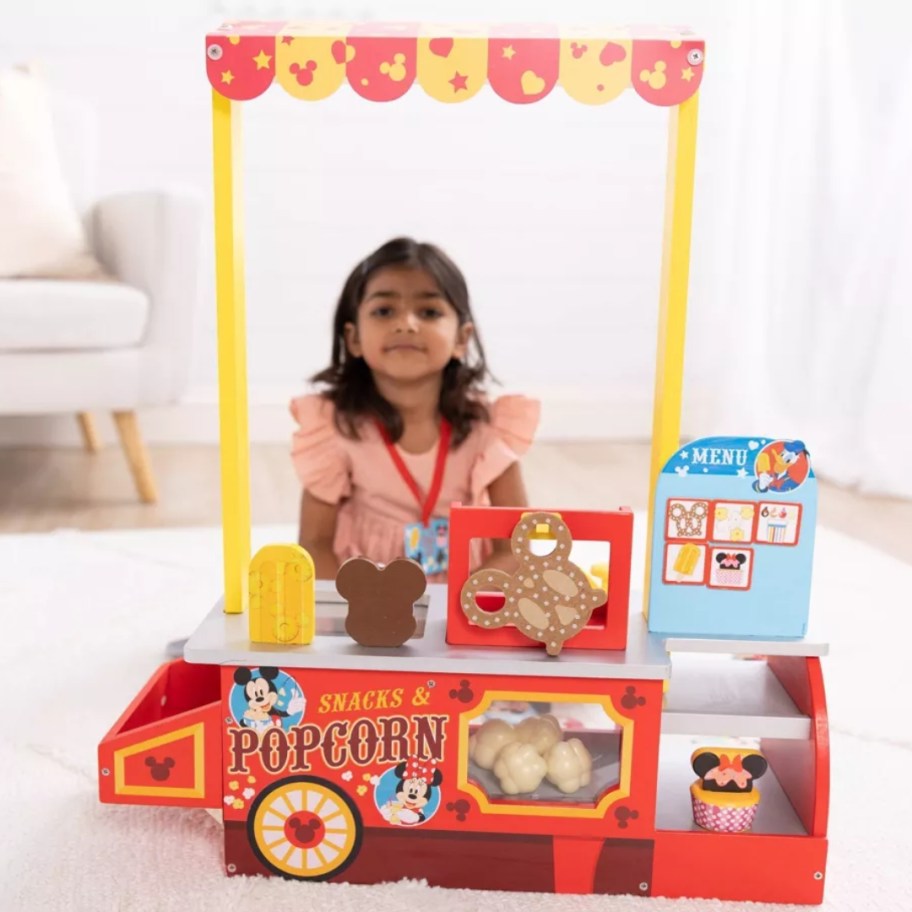 little girl sitting behind a toy popcorn and snacks food counter playset featuring Mickey & Minnie Mouse & other Disney characters.