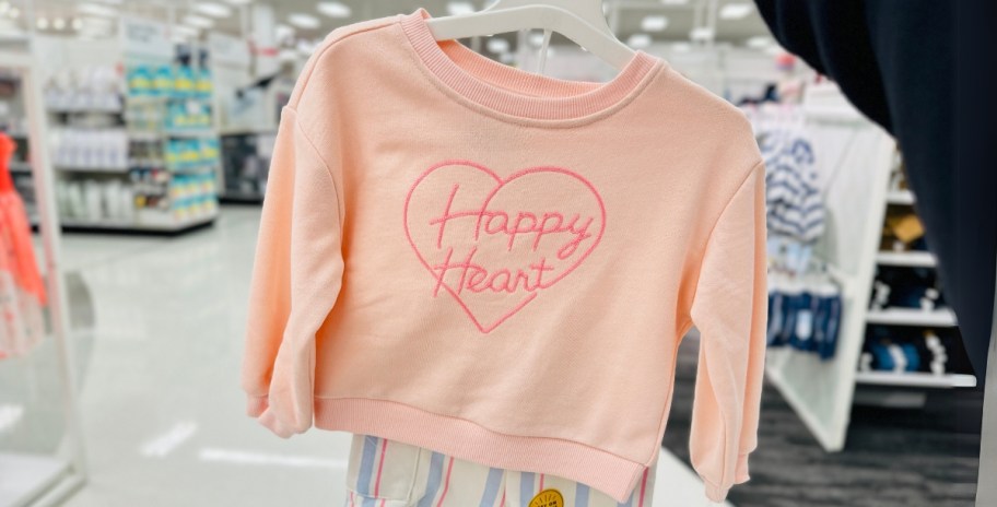 hand holding a girls valentines sweatshirt and shorts set on a hanger in Target, the sweatshirt is peach with a pink heart that says "Happy Heart" and the shorts are peach, pink, blue, and white striped