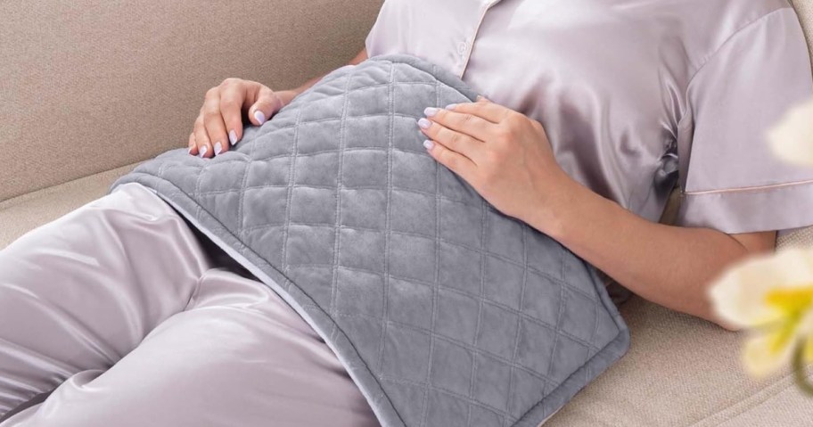 woman wearing button down light purple pajamas sitting on a tan couch with a grey quilted heating pad over her stomach