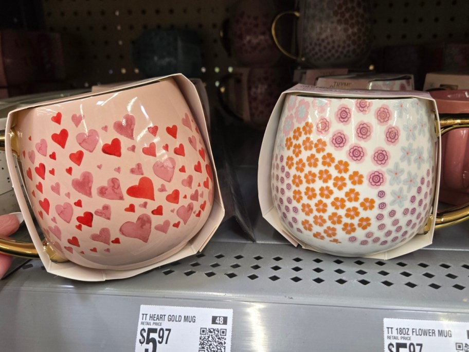 Two mugs with gold handles displayed on a store shelf. The left mug is pink with red and pink hearts, and the right mug is white with orange and purple flowers. Price tags are visible below.