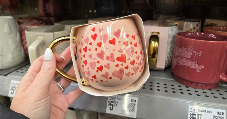 A pink mug with red heart patterns and a gold handle displayed in packaging. Price tag shows $5.97. Hand with white nail polish holds it.