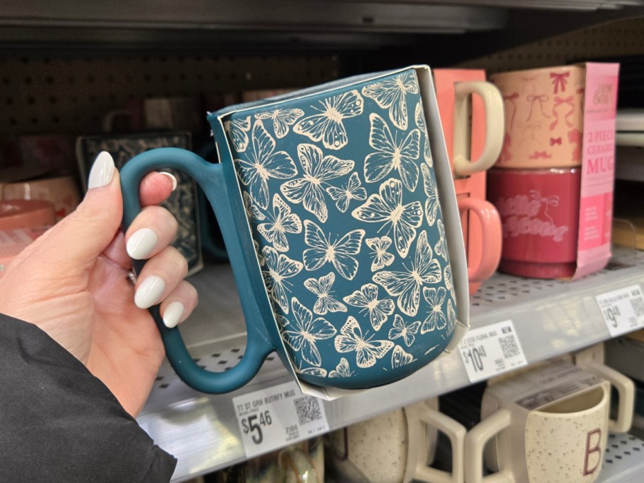A hand with white-painted nails holds a teal mug adorned with white butterflies, displayed on a store shelf.