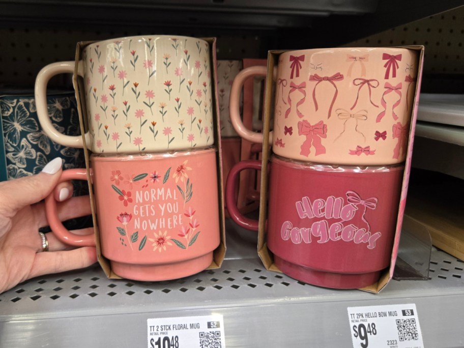 Two sets of stacked mugs on a store shelf. Left set: floral designs with "Normal gets you nowhere." Right set: bow designs with "Hello Gorgeous.