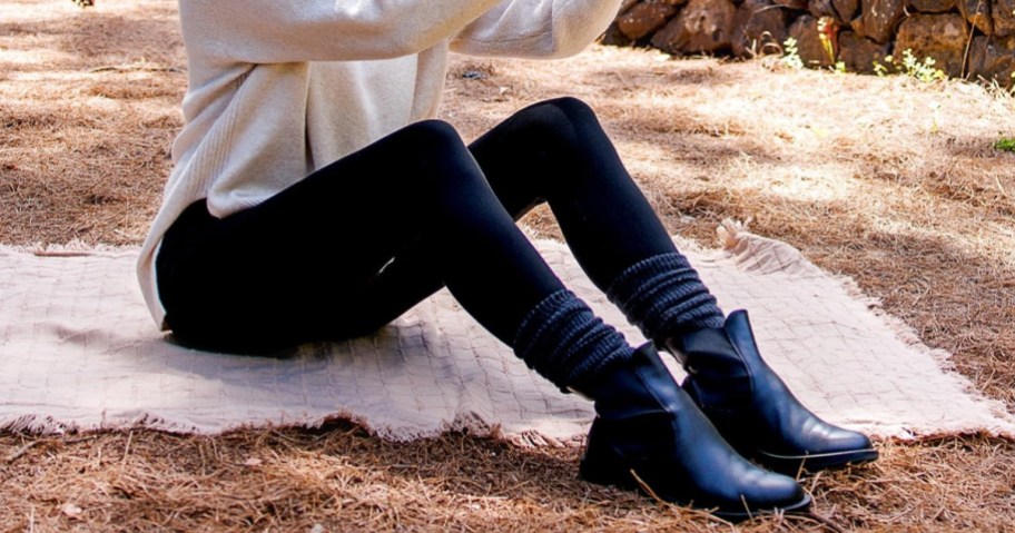 A person wearing black leggings, a light-colored sweater, and black boots sitting outdoors on a blanket spread over a bed of pine needles.
