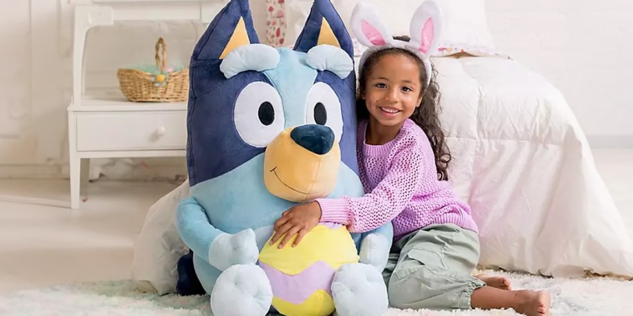 little girl sitting with large bluey plush in front of bed