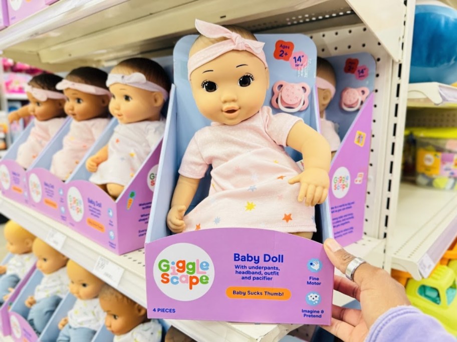 a baby doll with a white skin tone and brown eyes wearing a light pink dress with stars on it and a matching headband sitting in the packaging it comes in on a store shelf, more dolls behind it