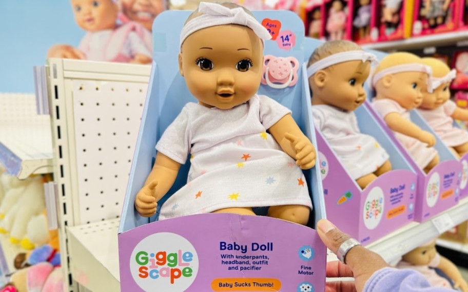 a baby doll with a light brown skin tone and brown eyes wearing a light pink dress with stars on it and a matching headband sitting in the packaging it comes in on a store shelf, more dolls behind it