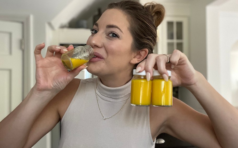 woman sipping on ginger shot holding up two other yellow juice shot glasses