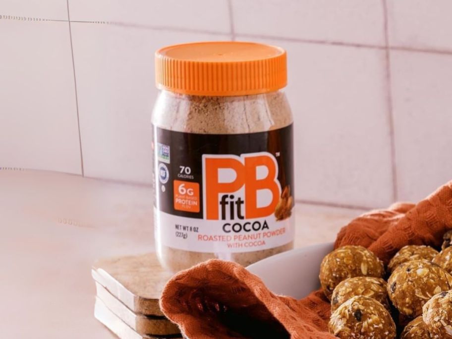 PBfit Cocoa Peanut Butter Powder 8oz container on counter next to energy balls