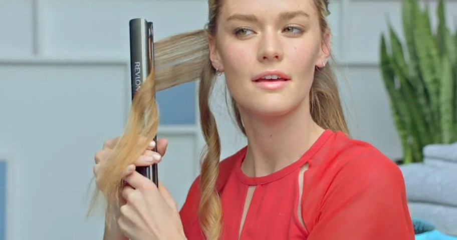 woman with long blonde hair wearing a red shirt, using a flat iron to curl her hair