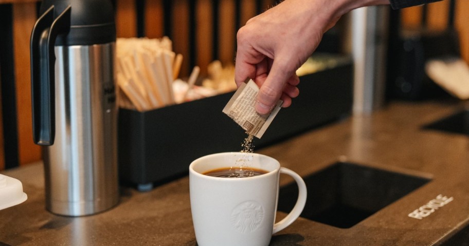 person pouring sugar from a paper packet into a white cup of coffee