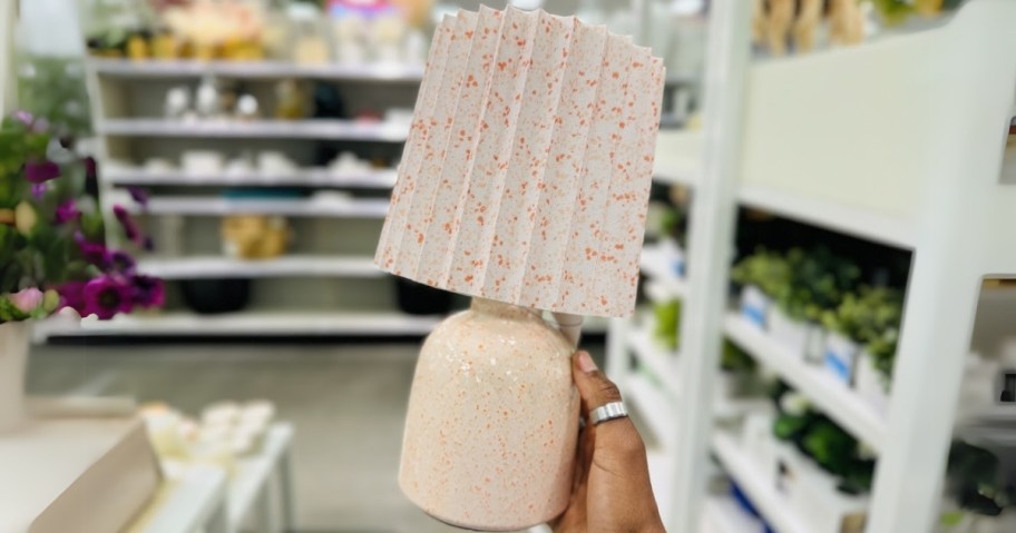 hand holding a pinkish peach color table lamp with a speckled pleated shade