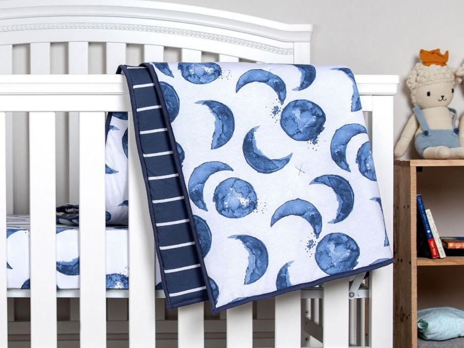 A crib with a Burt's Bees Hello Moon! Organic Cotton Reversible Baby Quilt hanging on it