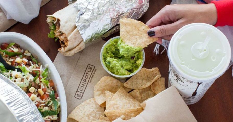 hand dipping a tortilla chip in a cup of guacamole on a chipotle napkin with a burrito next to it